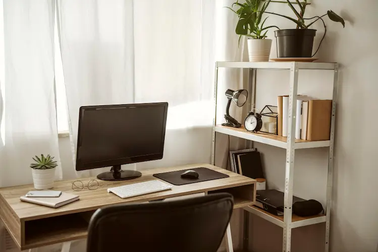 A modern home office with a wood desk, computer monitor, black chair, and a white shelving unit holding plants and books.