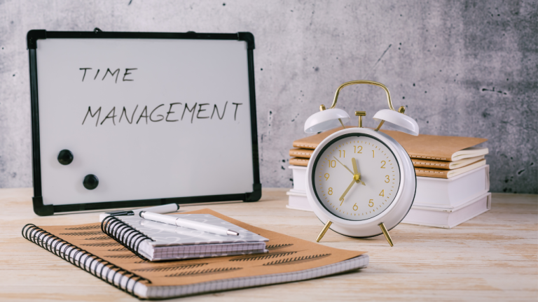 A time management concept on a light wood desk. On the left, a small whiteboard with a black frame displays the handwritten words 'TIME MANAGEMENT' in black marker. Beside it stands a white and gold retro-style twin-bell alarm clock showing approximately 8:05, resting on a stack of closed notebooks with brown and white covers. In the foreground, two spiral notebooks lie open—one with a brown cover featuring arrow patterns and a white pen resting on top. The background is a textured, light gray concrete wall.