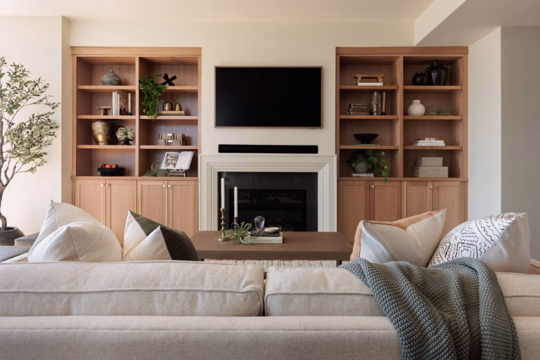 A cozy, modern living room featuring a large beige sectional sofa with assorted throw pillows and a chunky sage green knitted blanket draped over the back. In the center, a dark wood coffee table holds candles and a stack of books. The focal point is a white fireplace with a black insert, framed by light oak built-in cabinets and open shelves displaying curated decorative items and trailing plants. A wall-mounted TV is centered above the fireplace, and a potted olive tree stands to the left.