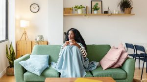A woman with long, dark curly hair, wearing a black-and-white striped long-sleeved shirt, sits on a green sofa holding a white mug with both hands and looking thoughtfully off-camera to the left. She has a light blue blanket draped over her lap and legs. A matching light blue throw pillow sits to her left, while two dusty pink throw pillows rest on her right. The room has a bright, cozy interior with light wood floors and a white wall. To the left, a wooden side cabinet holds a lamp with a tripod base and a trailing plant, with a potted snake plant in a woven basket on the floor. Above, a light wood floating shelf displays framed artwork, books, and small potted plants. A round black wall clock is mounted high on the left wall, and in the background on the right, two blue chairs are partially visible.