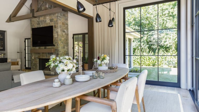 A bright dining area with an oval wooden table, white chairs, a stone fireplace, and large garden windows.