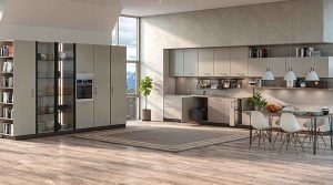 A bright, modern open-concept living space with light wood flooring and neutral gray cabinetry. On the left, a large floor-to-ceiling storage unit features an open bookcase, glass display sections, and a built-in oven. A tall window in the center offers a view of snowy mountains. To the right, a minimalist kitchen with sleek gray cabinets, a stainless steel sink, and a hanging utensil rack is next to a dining area with a wooden table, white molded chairs, and three pendant lights hanging overhead. A large rectangular area rug and a potted plant sit in the open floor space, illuminated by strong natural sunlight.