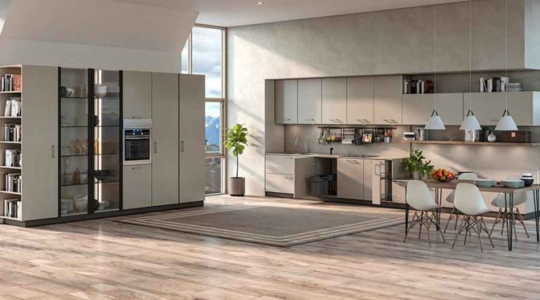 A bright, modern open-concept living space with light wood flooring and neutral gray cabinetry. On the left, a large floor-to-ceiling storage unit features an open bookcase, glass display sections, and a built-in oven. A tall window in the center offers a view of snowy mountains. To the right, a minimalist kitchen with sleek gray cabinets, a stainless steel sink, and a hanging utensil rack is next to a dining area with a wooden table, white molded chairs, and three pendant lights hanging overhead. A large rectangular area rug and a potted plant sit in the open floor space, illuminated by strong natural sunlight.