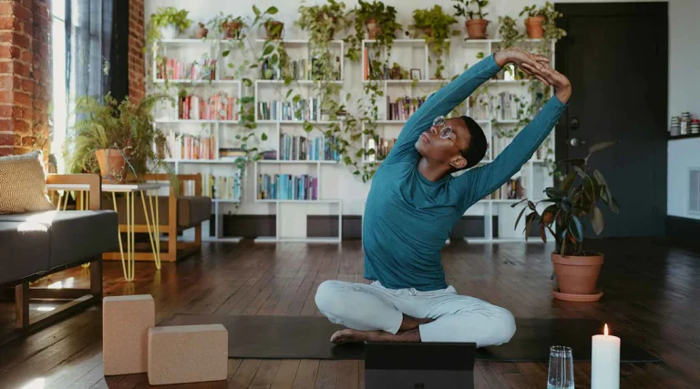 A person with short dark hair and glasses, wearing a teal long-sleeved top and white pants, sitting cross-legged on a black yoga mat on a dark hardwood floor. They are performing a seated side stretch with both arms raised overhead and clasped together, looking upward. To their left on the mat are two stacked cork yoga blocks. In front of them sits an open laptop, a glass of water, and a lit white candle. The room features an exposed brick wall on the left, a dark sofa, and a small side table with yellow hairpin legs. The background is dominated by a large, white, geometric bookshelf filled with books and lush trailing green houseplants. A large potted plant sits on the floor to the right, near a dark door.