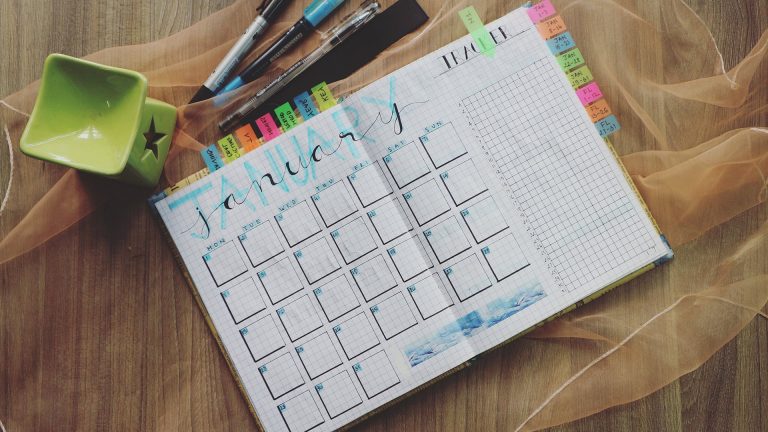 A flat-lay photograph of a hand-drawn bullet journal opened to a monthly calendar page for January, laid on a wooden surface atop a piece of sheer, peach-colored fabric. The page features a large, decorative 'JANUARY' header in black and light blue lettering, with a grid of hand-drawn boxes for the days of the month and a checklist section on the right. Several colorful sticky index tabs project from the top and right edges of the page. Three pens, a black marker, and a bright green, star-shaped ceramic container sit on the left side of the journal.