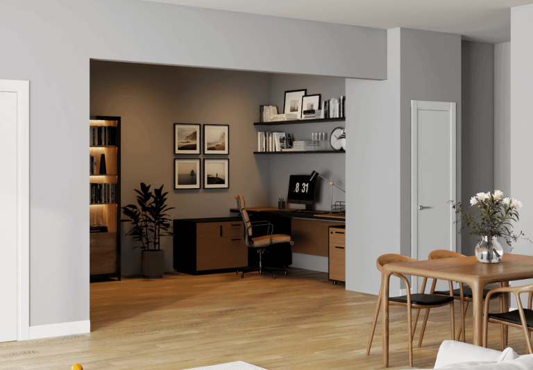 A modern, open-concept interior featuring a built-in home office nook recessed into a light gray wall. The office space is painted in a warm taupe tone and contains a sleek dark wood L-shaped desk holding a computer monitor displaying the time '8:31', a silver desk lamp, and a black filing cabinet with drawers. A tan leather office chair with a chrome base sits at the desk. Above the desk, two floating black shelves display books and a round wall clock. On the left wall of the nook, four framed black-and-white landscape photographs are arranged in a square grid. To the far left, a tall black bookcase with warm internal lighting holds books and bottles, and a tall potted plant stands nearby. In the foreground on the right, a light wood dining table with matching chairs is topped with a clear glass vase containing white flowers. The flooring throughout is light wood, and a white door is visible on the far right.