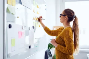 A woman with long brown hair tied in a high ponytail, wearing black-rimmed glasses and a mustard-yellow textured knit sweater, standing in profile facing a large whiteboard. She is holding a white marker in her right hand and writing on a cluster of yellow and pale green sticky notes arranged on the board. Several printed papers and additional notes are attached to the board with magnets below. The background is a bright, softly lit room with a plant visible in the lower corner.