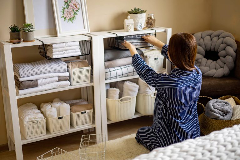 A woman with short brown hair, wearing navy blue and white striped pajamas, kneeling on a fluffy white rug in a cozy, organized room. She is reaching up with both hands to slide a black wire drawer into a white shelving unit, organizing her linens. The two white wooden shelves are neatly arranged with folded towels, stacked blankets, and white woven storage baskets. On the top shelf, framed botanical artwork, small potted succulents, a white ceramic jar, and a tiny clock are displayed. To the right, a dark sofa features a large, chunky gray knot pillow. In the foreground, a white knit blanket is partially visible, along with a brown woven basket holding rolled towels and empty wire baskets on the floor.