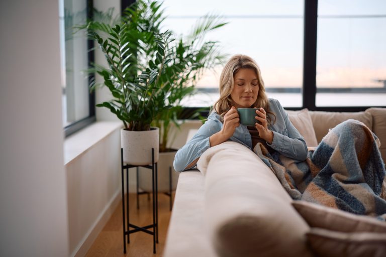 A woman with blonde hair wearing a light blue denim shirt sits comfortably on a cream-colored sofa, holding a small teal mug with both hands and looking down at it with a peaceful, relaxed expression. Her legs are covered by a thick plaid blanket featuring wide stripes of blue, tan, and brown. To her left, a large, leafy green houseplant sits in a textured white pot elevated on a tall, black metal plant stand. The background consists of large, black-framed windows offering a view of a tranquil sky and water at what appears to be sunrise or sunset, filling the space with soft, warm light.