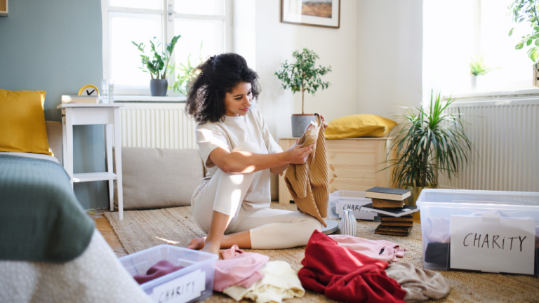 A young woman with curly dark hair, wearing a cream-colored t-shirt and leggings, sitting cross-legged on a woven rug in a bright, sunlit room. She is holding up a brown garment while sorting through a pile of clothing, with stacks of books and several clear plastic storage bins labeled 'CHARITY' resting nearby on the floor. Potted green plants, a white desk with a clock, and a window fill the cozy background.