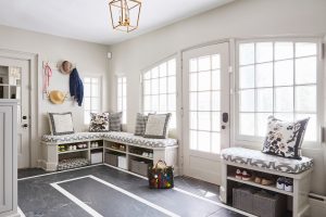A bright, well-organized mudroom with light gray walls and a dark, polished floor accented by a large white geometric border. A built-in corner bench with white cabinetry runs along the walls, topped with grey-and-white patterned cushions and a collection of decorative throw pillows, including floral and black-and-white houndstooth designs. Underneath the benches, open cubbies hold neatly stored shoes and grey woven storage baskets. A small, separate bench on the right also features a floral pillow and storage for shoes and a basket below. On the left wall, a brass coat rack with gold hooks holds a straw hat, a dark blue jacket, and a pink leash. A large, multi-colored striped tote bag sits on the floor in the center. A gold geometric lantern pendant light hangs from the ceiling, and light streams through large white French doors and multi-paned windows.