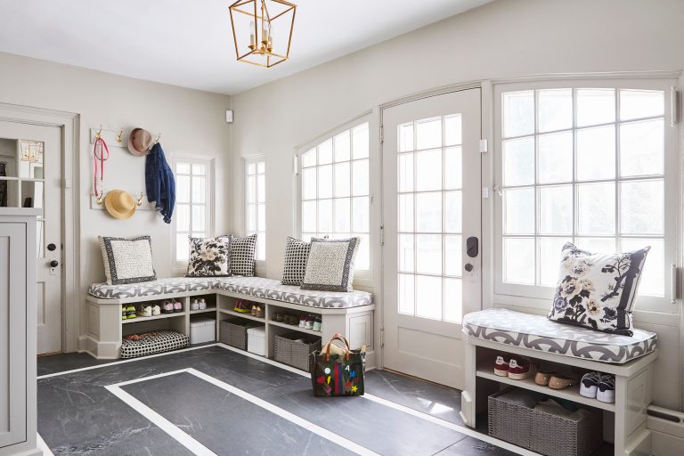 A bright, well-organized mudroom with light gray walls and a dark, polished floor accented by a large white geometric border. A built-in corner bench with white cabinetry runs along the walls, topped with grey-and-white patterned cushions and a collection of decorative throw pillows, including floral and black-and-white houndstooth designs. Underneath the benches, open cubbies hold neatly stored shoes and grey woven storage baskets. A small, separate bench on the right also features a floral pillow and storage for shoes and a basket below. On the left wall, a brass coat rack with gold hooks holds a straw hat, a dark blue jacket, and a pink leash. A large, multi-colored striped tote bag sits on the floor in the center. A gold geometric lantern pendant light hangs from the ceiling, and light streams through large white French doors and multi-paned windows.