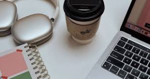 A flat-lay view of a white desk surface featuring a silver laptop with a black keyboard, a white spiral notebook with a geometric pink and green cover, a white and silver pair of headphones in their charging case, and a paper coffee cup with a black lid and a brown corrugated sleeve.