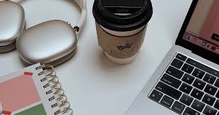 A flat-lay view of a white desk surface featuring a silver laptop with a black keyboard, a white spiral notebook with a geometric pink and green cover, a white and silver pair of headphones in their charging case, and a paper coffee cup with a black lid and a brown corrugated sleeve.