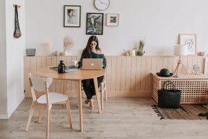 A woman with long dark hair and glasses, wearing a dark green off-the-shoulder top and black pants, sits barefoot at a round, light-wood dining table working on a silver laptop. A white chair with wooden legs is placed at the table, along with a dark bottle and an open magazine. The room has a minimalist, Scandinavian-inspired aesthetic with white walls, light wood flooring, and a horizontal wooden wainscoting ledge. Above the ledge, framed black-and-white photographs and a round wall clock are displayed. A woven macrame hanging and a black wire basket adorn the wall to the left. On the right, a wooden slatted storage bench holds a black bag and a woven hat, with a floor lamp and framed line-art print resting nearby, and a textured dark rug partially covering the floor.