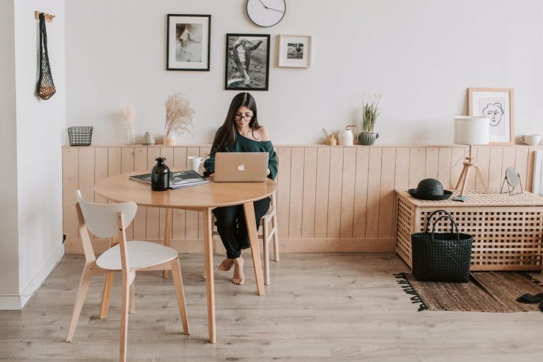 A woman with long dark hair and glasses, wearing a dark green off-the-shoulder top and black pants, sits barefoot at a round, light-wood dining table working on a silver laptop. A white chair with wooden legs is placed at the table, along with a dark bottle and an open magazine. The room has a minimalist, Scandinavian-inspired aesthetic with white walls, light wood flooring, and a horizontal wooden wainscoting ledge. Above the ledge, framed black-and-white photographs and a round wall clock are displayed. A woven macrame hanging and a black wire basket adorn the wall to the left. On the right, a wooden slatted storage bench holds a black bag and a woven hat, with a floor lamp and framed line-art print resting nearby, and a textured dark rug partially covering the floor.