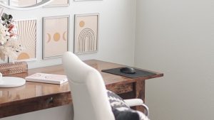 A cozy, neutral-toned workspace featuring a dark wood desk with a glossy finish. On the desk sits a white office chair, a black mousepad with a black computer mouse, and a white notebook. The wall is decorated with three framed art prints in soft beige and peach tones featuring bohemian-style designs: a sunburst, moon phases, and an arch with a circle. To the left, a decorative arrangement of dried flowers and pampas grass in a woven basket adds texture, with a round mirror partially visible above the frames.