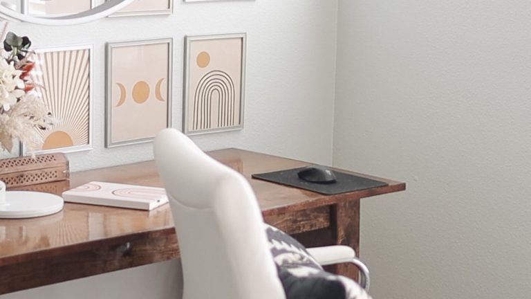 A cozy, neutral-toned workspace featuring a dark wood desk with a glossy finish. On the desk sits a white office chair, a black mousepad with a black computer mouse, and a white notebook. The wall is decorated with three framed art prints in soft beige and peach tones featuring bohemian-style designs: a sunburst, moon phases, and an arch with a circle. To the left, a decorative arrangement of dried flowers and pampas grass in a woven basket adds texture, with a round mirror partially visible above the frames.
