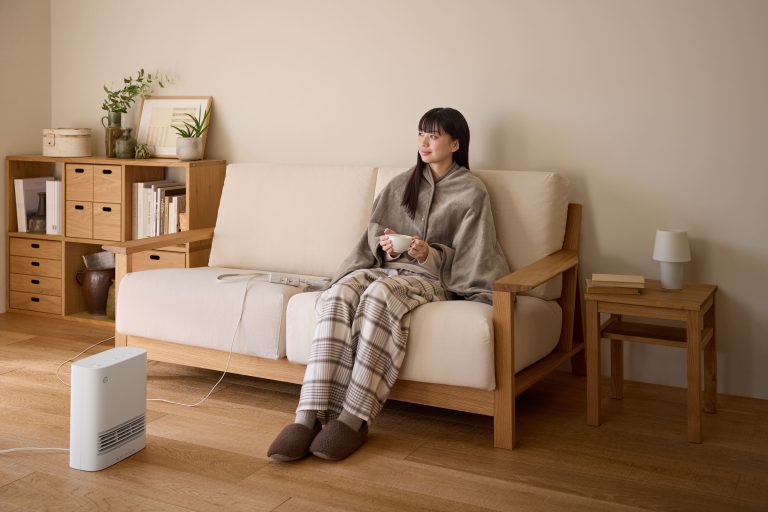A woman in cozy pajamas and a poncho sitting on a sofa with a mug, next to a space heater in a minimalist living room.
