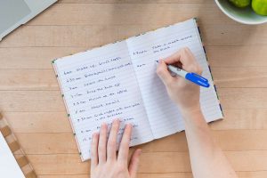 A person writing a handwritten daily schedule in an open notebook with a blue pen.