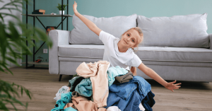A young girl with her hair tied back, wearing a white t-shirt, kneeling on a light wood floor behind a large, messy pile of folded and unfolded laundry. She looks directly at the camera with a slight smile, posing playfully with both arms stretched out wide in a celebratory or triumphant gesture. A light gray sofa with pillows sits behind her against a soft teal wall, with a black metal shelving unit holding small plants and books in the background, and blurred green foliage in the left foreground.