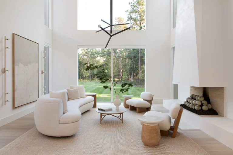 A spacious, double-height modern living room with a bright, minimalist aesthetic and expansive views of a lush green lawn and forest through floor-to-ceiling glass windows. The room features a large, curved white sectional sofa and matching white lounge chairs with rounded, wooden bases, arranged around a small round coffee table that holds a book and a white ceramic vase with a leafy branch. A textured, light beige rug anchors the seating area. On the left wall hangs a large, abstract framed painting, illuminated by a modern, multi-bulb vertical wall sconce. A striking black, angled linear chandelier is suspended from the high white ceiling. On the right, a massive, white sculptural fireplace with a wide hearth displays a neatly arranged pile of round, gray stone fireballs. The overall atmosphere is serene, airy, and bathed in natural light.