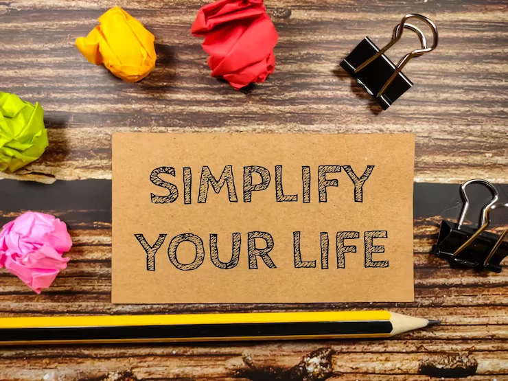 A flat-lay photograph on a dark, rustic wood-grain surface featuring a rectangular piece of brown kraft paper in the center, with the words 'SIMPLIFY YOUR LIFE' hand-drawn in bold, textured black sketch-style lettering. Surrounding the card are scattered office supplies: three crumpled balls of paper in yellow, red, and bright green at the top left; a crumpled pink paper ball at the bottom left; two black metal binder clips on the right side; and a yellow-and-black striped pencil lying horizontally across the bottom edge of the frame.