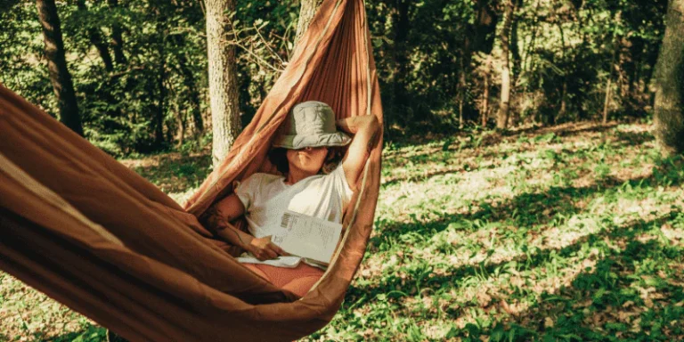 A person relaxing in a rust-orange fabric hammock suspended between trees in a sunlit woodland clearing. They are wearing a white t-shirt, reddish shorts, and a light-colored bucket hat pulled low over their face, and they are holding an open book resting on their lap while one arm is tucked comfortably behind their head. The lush green forest floor is covered in dappled sunlight and scattered leaves, creating a tranquil, summery outdoor atmosphere.