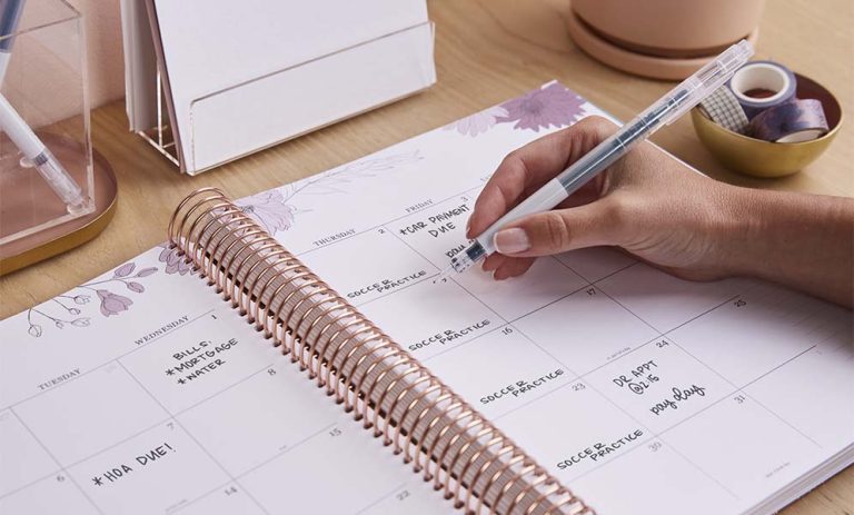 A close-up view of a person's hand writing in an open, spiral-bound monthly planner with a rose-gold coil and light purple floral illustrations along the margins. The person is holding a white pen with a clear barrel and a blue grip, writing on a day's calendar box. The planner page shows pre-written reminders and appointments, including 'CAR PAYMENT DUE' on Friday the 3rd, 'BILLS MORTGAGE WATER' on Wednesday the 1st, 'HOA DUE!' on Tuesday the 6th, 'SOCCER PRACTICE' on several days, and 'DR APPT @ 2:15 my day off' later in the month. The planner rests on a light wood desk. To the left, a clear acrylic pen holder and a white document organizer are partially visible. To the right, a small gold bowl holds rolls of decorative washi tape, and a pink planter sits in the softly blurred background.