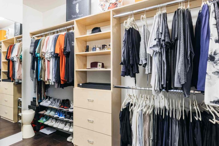 A brightly lit, organized walk-in closet featuring light wood cabinetry and two main hanging sections. The left side holds a colorful assortment of short-sleeved shirts and polos, with a multi-tiered shoe rack below displaying sneakers. The center features a column of drawers and open shelves holding men's cologne bottles and a small framed photo. The right side has two hanging rods: the top holds casual t-shirts and hoodies in shades of gray, black, and navy, while the bottom displays neatly hung pants and trousers on white hangers. A tall mirror and a white stool are visible on the far left.