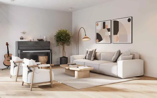 A bright, minimalist living room featuring a modern and musical aesthetic. On the right, a large, light beige modular sofa is styled with grey and patterned throw pillows, sitting against a white wall adorned with two large framed abstract art prints in earthy tones of beige, black, and peach. A sleek floor lamp with an arched stem and a brass shade provides accent lighting near the art. In the center, a light wood, geometric low coffee table rests on a large textured beige area rug, holding a couple of books. Two cream-colored armchairs with light wood frames are positioned facing the table on the left. Behind them, against the back wall, sits a dark grey upright piano with a few small decor items resting on top, and an acoustic guitar stands on a floor stand to its left. A tall, bushy green potted plant stands between the piano and the sofa, adding a touch of nature. The room features light wood flooring, recessed ceiling lights, and soft natural light creating gentle shadows.