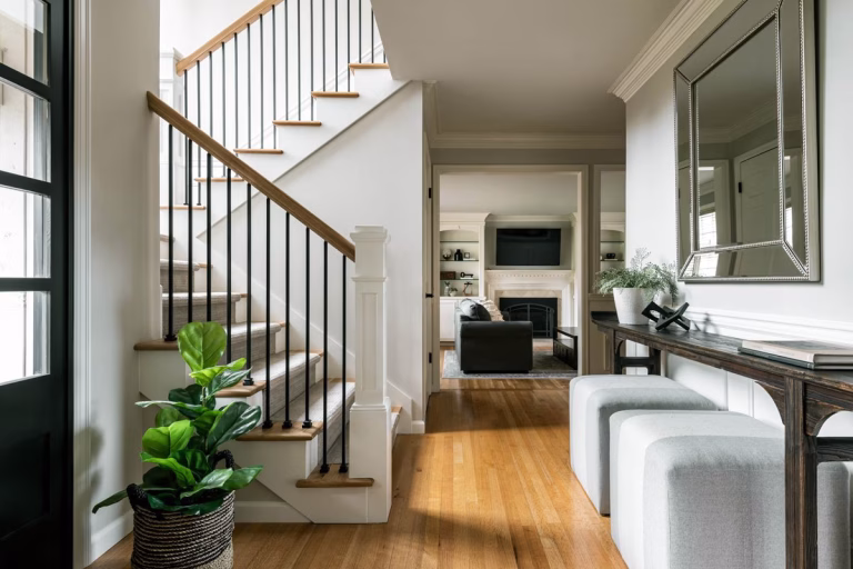 A bright, modern entryway featuring a staircase on the left with white risers, light oak wood treads, and a railing composed of thin black metal balusters and a matching wood handrail. A large, vibrant green potted plant sits on the floor in a woven basket at the base of the stairs. On the right, a long, dark wood console table runs along the white wall, holding a small potted plant and a decorative black book stand. Above the table hangs a large rectangular mirror with a beaded silver frame. Tucked underneath the console are two light gray, cube-shaped upholstered ottomans. The floor is polished light hardwood. Through an open doorway in the center, the view extends into a cozy living room featuring a gray sofa, a white fireplace mantel with a black firebox, a flat-screen television, and built-in white shelving on either side.