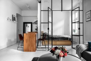 A bright, contemporary studio apartment featuring a striking industrial-style room divider made of black metal frames and glass panes, which separates the living space from the bedroom area. Through the glass partition, a bedroom is visible with a low wooden platform bed, a gray duvet, and a window with a small potted plant. In the center foreground, a wooden kitchen island counter stands with two brown leather bar stools on slim black legs. A stainless steel range hood is visible above the counter in the background. In the immediate foreground, a round, dark coffee table holds a white vase filled with a vibrant bouquet of pink, red, white, and dark purple flowers. To the right, a gray sofa is partially visible, draped with a textured light gray throw blanket and a dark teal pillow. White walls, light flooring, and a picture shelf with a framed photo and decorative object complete the bright, minimalist interior.