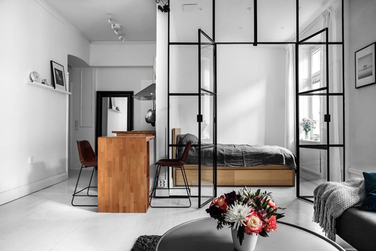 A bright, contemporary studio apartment featuring a striking industrial-style room divider made of black metal frames and glass panes, which separates the living space from the bedroom area. Through the glass partition, a bedroom is visible with a low wooden platform bed, a gray duvet, and a window with a small potted plant. In the center foreground, a wooden kitchen island counter stands with two brown leather bar stools on slim black legs. A stainless steel range hood is visible above the counter in the background. In the immediate foreground, a round, dark coffee table holds a white vase filled with a vibrant bouquet of pink, red, white, and dark purple flowers. To the right, a gray sofa is partially visible, draped with a textured light gray throw blanket and a dark teal pillow. White walls, light flooring, and a picture shelf with a framed photo and decorative object complete the bright, minimalist interior.