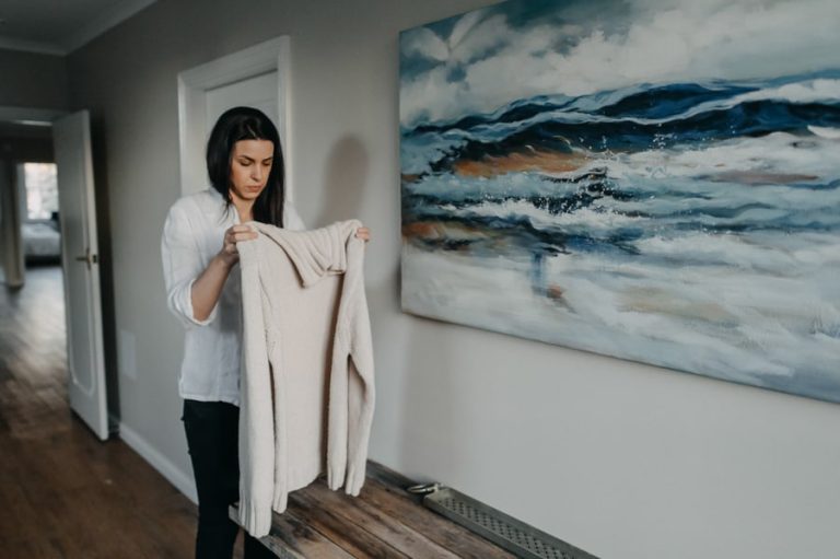 A woman with long dark hair, wearing a white button-down shirt and dark pants, stands in a hallway holding up a cream-colored knit cardigan with both hands, looking at it thoughtfully as if examining or organizing it. A white door frame is visible directly behind her, and an open doorway to the left reveals a glimpse into another room with a bed. To her right, a large, framed abstract painting depicting a stormy seascape in shades of deep blue, grey, and white hangs on the light-colored wall. Beneath the painting, a rustic wooden console table or bench rests against the wall, with a small white object near its edge. The room has a warm, natural aesthetic with dark hardwood flooring.