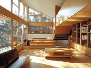 A striking, sun-drenched modern living space characterized by expansive, custom-designed light wood cabinetry and architectural details. The room features soaring, angled ceilings with massive, multi-pane windows that frame a view of tall pine trees outside. The interior is dominated by warm, honey-toned wood, including a large built-in bookcase that spans the entire right wall, filled with books and decorative objects. In the center of the space, a unique, low-slung geometric wooden coffee table rests on the polished wood floor. In the background, a tiered wooden platform or step structure leads to a higher level, suggesting a split-level layout. A dark, leather-like sofa sits in the lower-left foreground, partially in shadow. Sunlight streams intensely through the windows, casting bright, geometric shadows across the floor and walls, creating a serene, organic, and contemporary atmosphere.